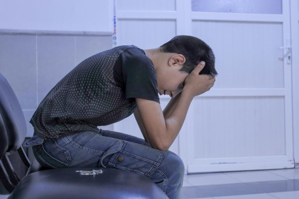 A young boy shows distress while sitting alone in a hospital waiting room, head in hands, conveying emotions of anxiety and sadness.
